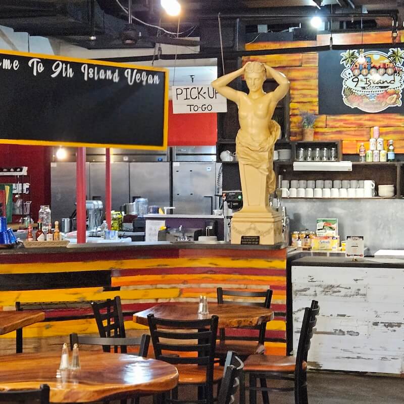 Cafe interior with wooden tables, a statue on the counter, and signs for "9th Island Vegan" and "Pick-Up To-Go" amid tropical decor.