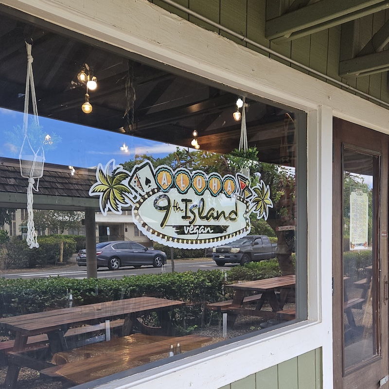 Exterior window of 9th Island Vegan restaurant with a sign reading 'Aloha 9th Island Vegan,' reflecting outdoor seating and nearby street