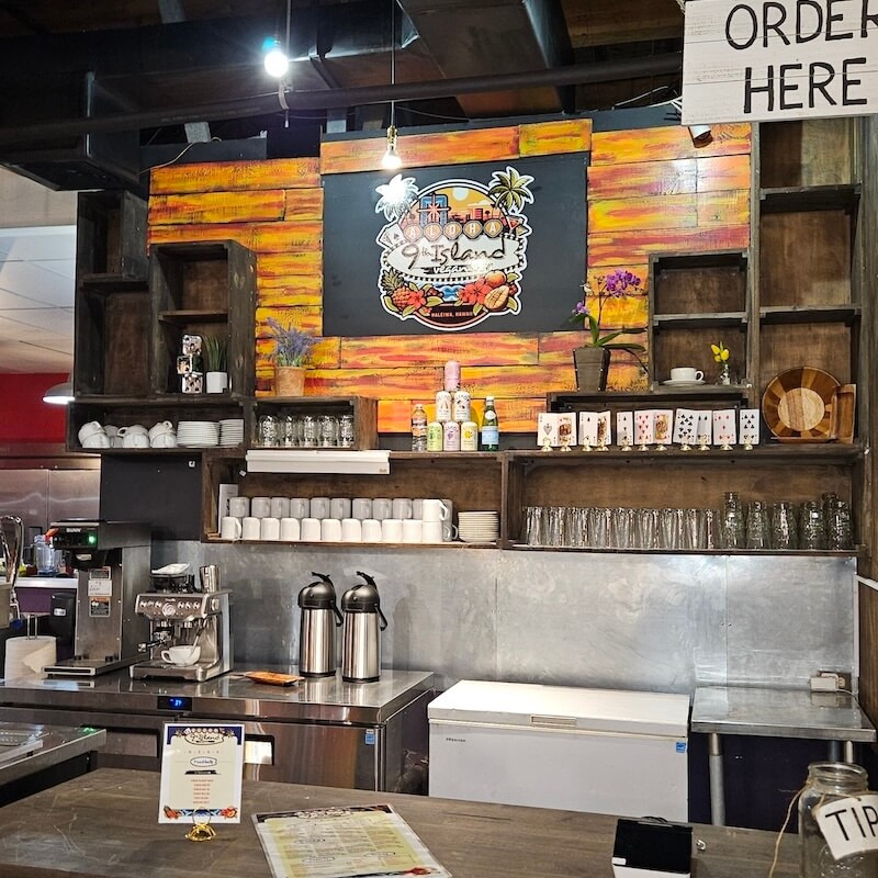 Cafe counter with tropical decor, rustic wooden shelves, a coffee machine, and a colorful island-themed logo on the wall.